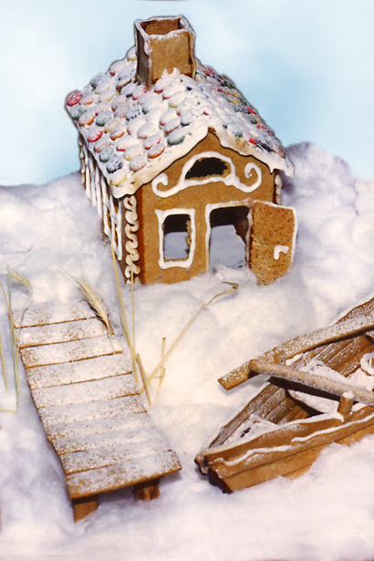 Foto av pepparkakshusi form av ett litet hus och framför det en sned och vind träbrygga som det ligger en gammal eka vid, med årorna lämnade i båten